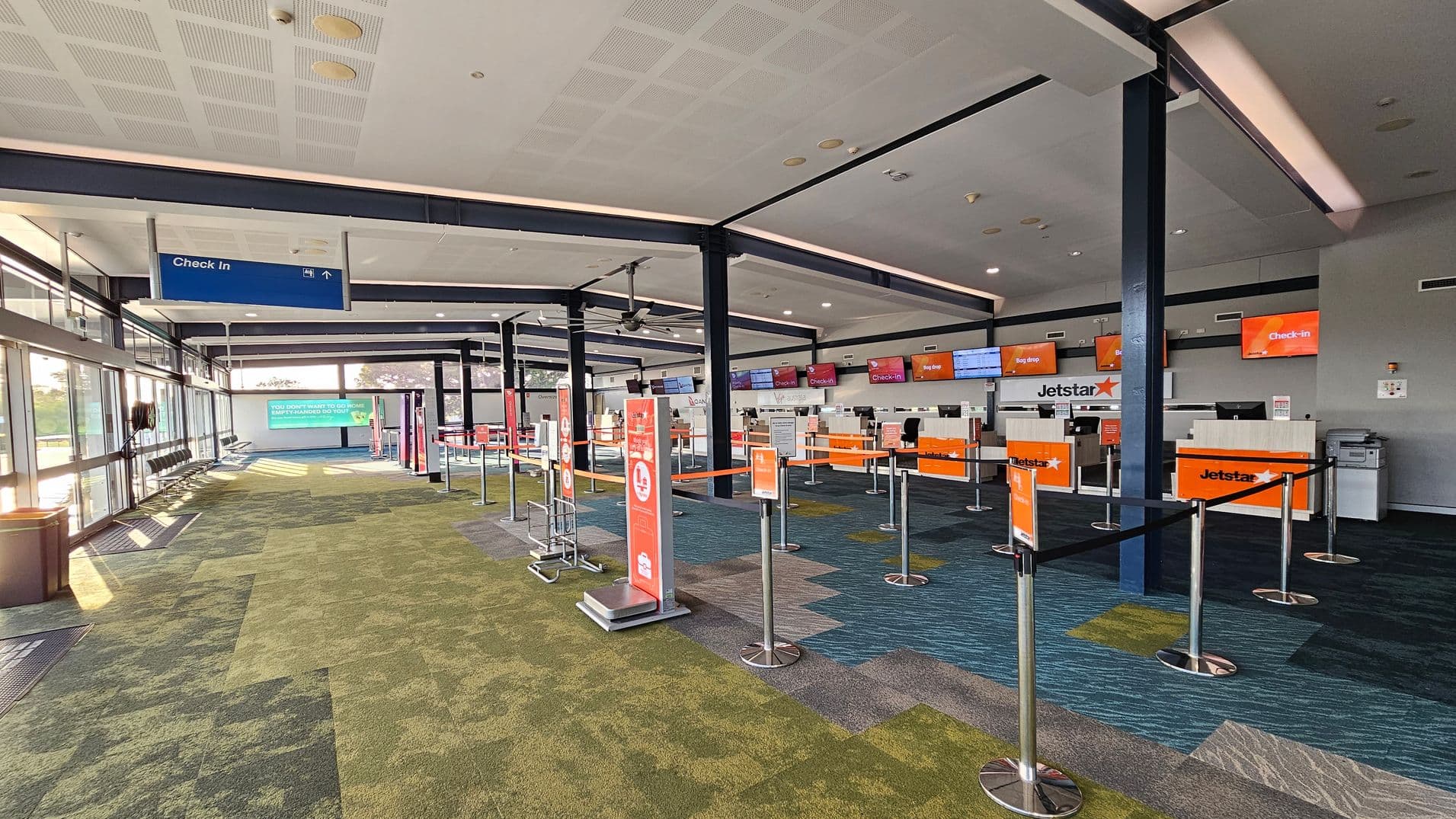An airport check-in area with multiple counters on the right, each displaying orange signs with "Jetstar" and "Check-in". The counters are separated by retractable barriers. On the left, there are large windows allowing natural light in, with a row of seats and a blue "Check In" sign hanging from the ceiling. The floor is covered with a patterned carpet in shades of green and blue. The ceiling is white with recessed lighting.