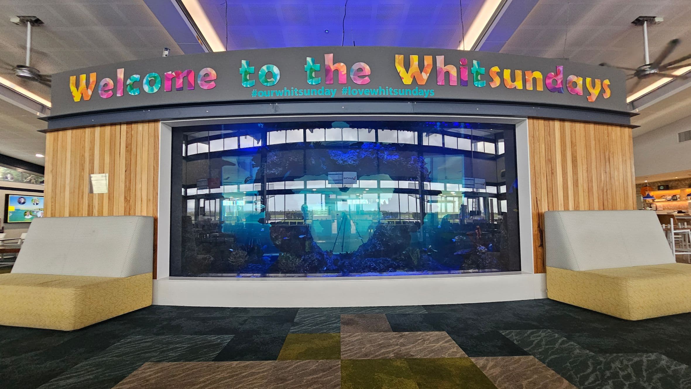 A large aquarium is set into a wooden panelled wall with colourful letters above spelling "Welcome to the Whitsundays" and hashtags below. On either side of the aquarium are cushioned seats with light green backs and beige bases. The floor is carpeted in a patchwork of dark and light grey patterns. The ceiling is grey with recessed lighting and ceiling fans. In the background, there are tables and chairs, suggesting a café or waiting area.