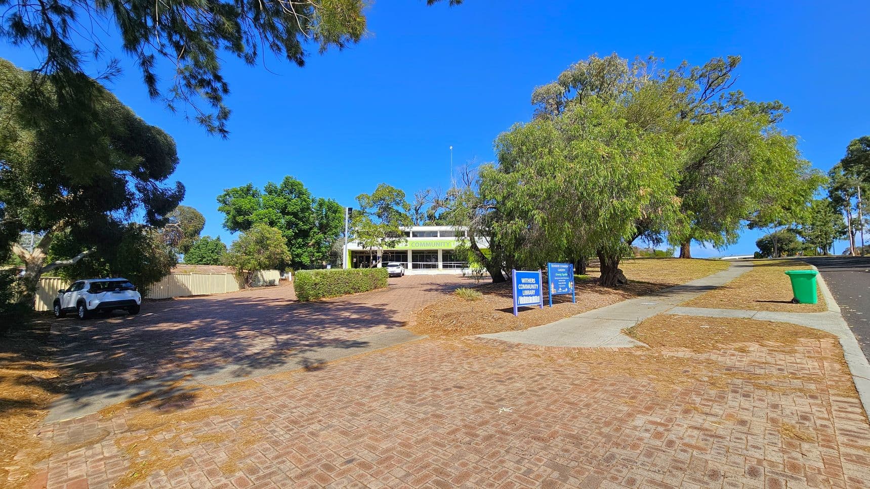 A paved pathway leads to a building in the centre, partially obscured by trees. On the left, a white car is parked near a beige fence, surrounded by trees. On the right, a green bin is placed on a concrete path, with more trees and a grassy area. Two blue signs are visible near the building, which has a sign reading "COMMUNITY" above the entrance. The ground is covered with fallen leaves and the sky is clear.