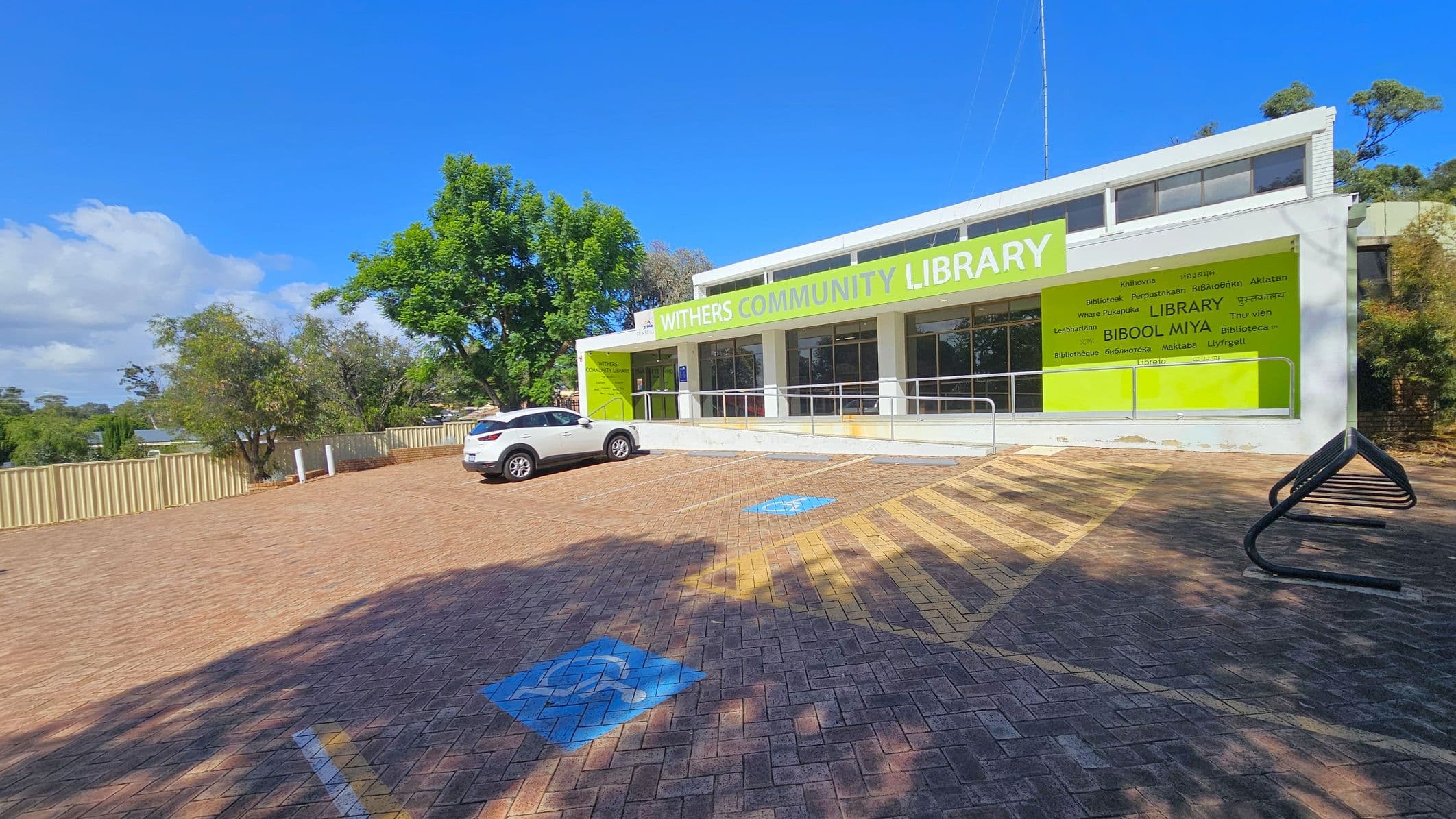 A single-storey building with a large green sign reading "Withers Community Library" on the right. The building has a white facade with multiple windows. In front, there is a paved parking area with a white car parked on the left. Blue disabled parking symbols are visible on the ground. A metal bike rack is on the right. Trees and a beige fence line the background, with a clear blue sky above.