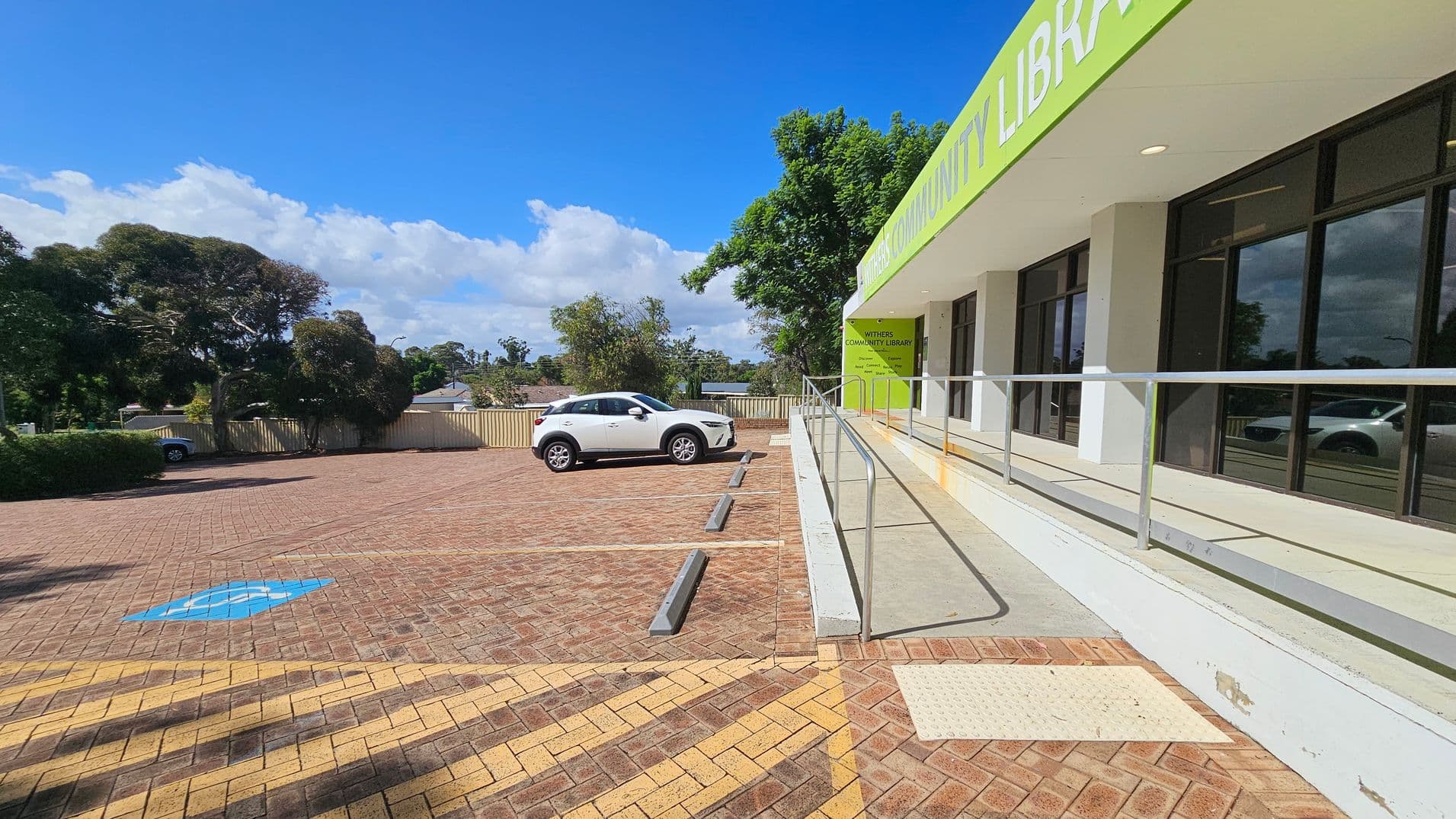 A parking area with brick paving is on the left, featuring a white car parked in a designated space. A blue disabled parking symbol is visible on the ground. On the right, a building with large windows and a green sign reading "Withers Community Library" is present. A ramp with a metal railing leads to the entrance. Trees and a fence are in the background, providing a natural setting.