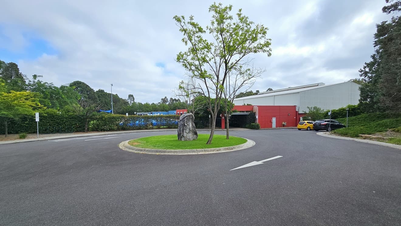 The road to Wollondilly Community Centre is divided by a traffic median, featuring a traffic sign. On the right stands a large banner displaying the facility name in the grass area. To the left is a green corrugated wall, surrounded by well-maintained grass and trees on both sides. Further ahead, cars are parked on both sides of the road.