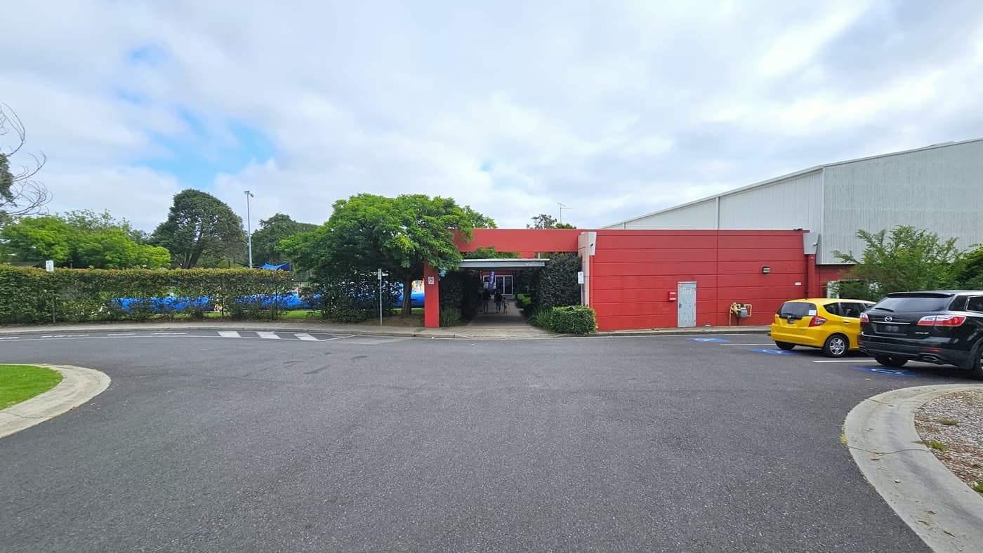The facility comprises a large white corrugated iron warehouse design with an adjoining red concrete building. Next to the building is the car park, featuring accessible slots marked with a blue wheelchair sign. Yellow and black cars are parked, surrounded by tall trees. Additional parking is available on the left, and a step-free ramp leads to a lengthy open-air corridor.