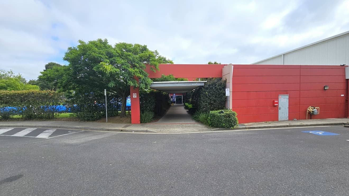The facility comprises a large white corrugated iron warehouse design with an adjoining red concrete building. Next to the building is the car park, featuring accessible slots marked with a blue wheelchair sign. Yellow and black cars are parked, surrounded by tall trees. Additional parking is available on the left, and a step-free ramp leads to a lengthy open-air corridor.