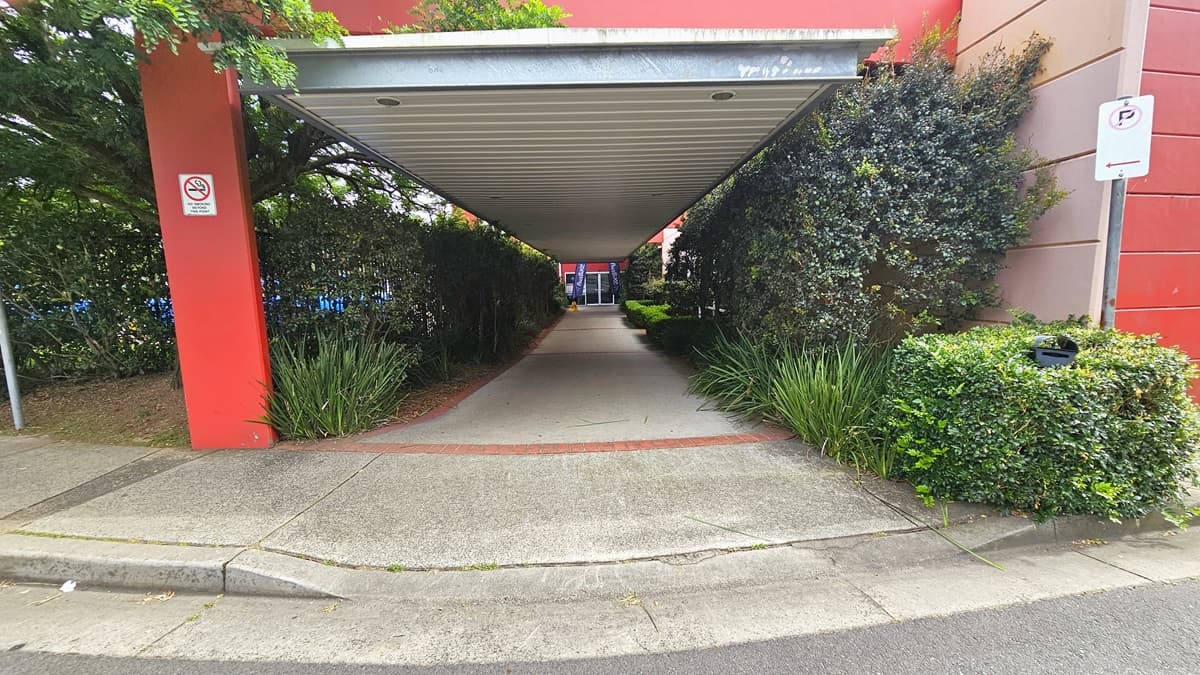 A step-free ramp leads to a long open-air corridor with a corrugated iron roof and a steel fence on the left. The pathway is adorned with trees, vines, and neatly trimmed shrubs. A "No Smoking" sign and a "Parking" sign are affixed to the post.