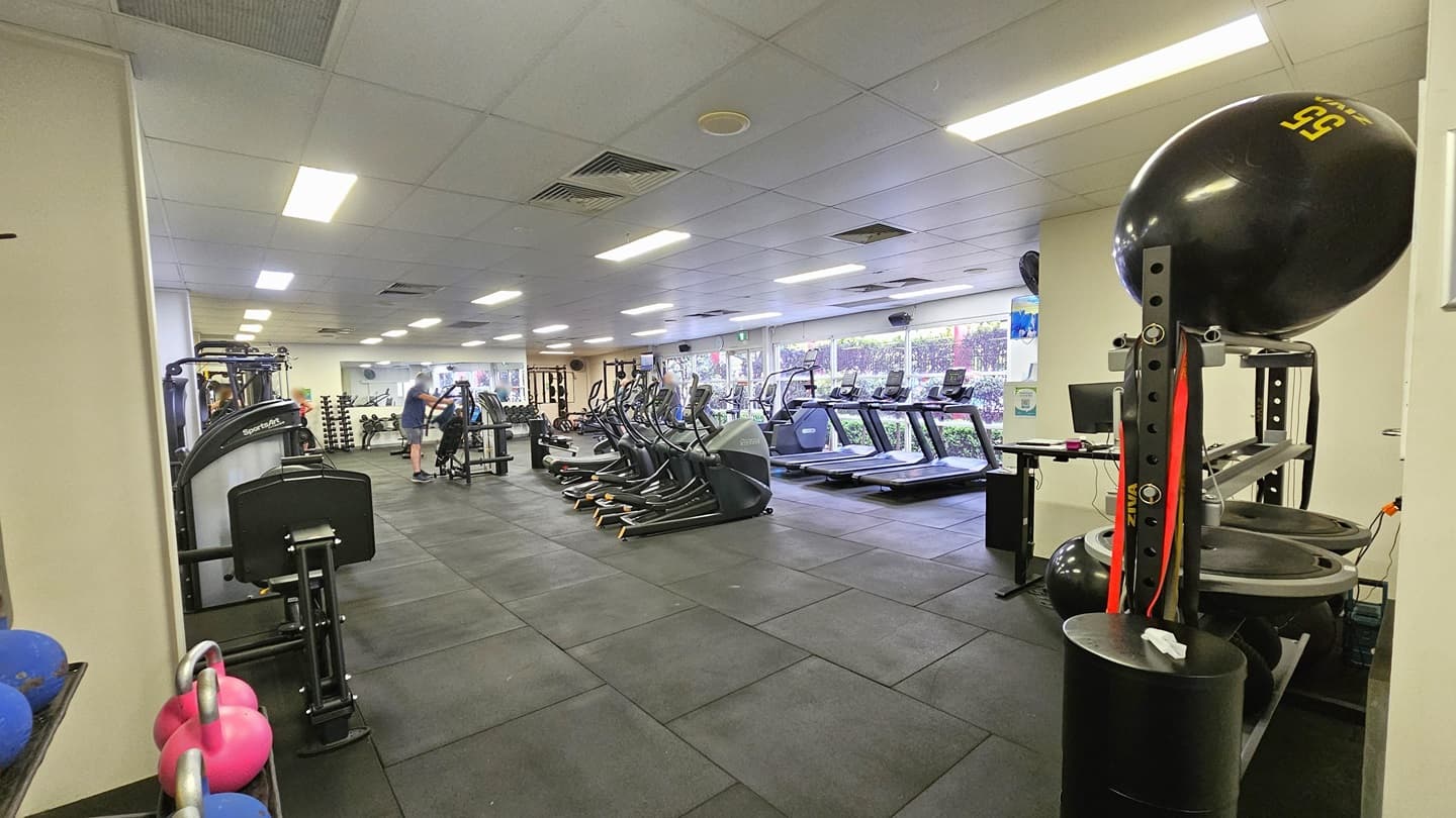 An outward-opening manual glass door is equipped with posters. A water dispenser is positioned on the door before entry. Inside, a white storage rack for belongings is placed on the right. Kettlebells are arranged on the left, near the wall. The floor features black rubber tiles, and the ceiling is all white with fluorescent lights.