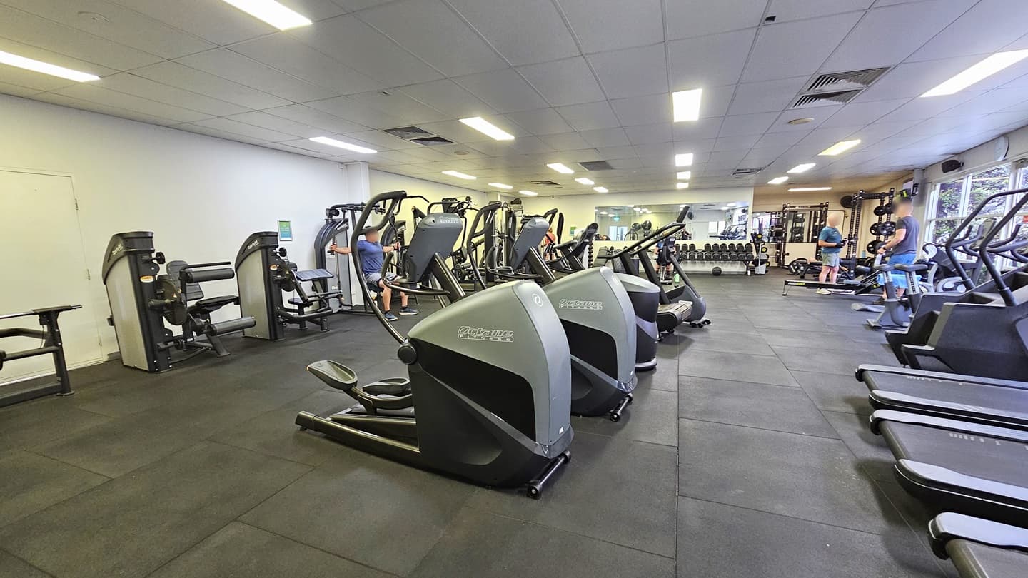 Treadmills and stairmills are positioned near the glass wall on the right. Lateral machines are lined up in the middle, and more strength machines are arranged on the left. Further in is the weights section where Smith Machines and free weights are arranged. The floor features black rubber tiles, and the ceiling is all white with fluorescent lights. Gym users are visible.