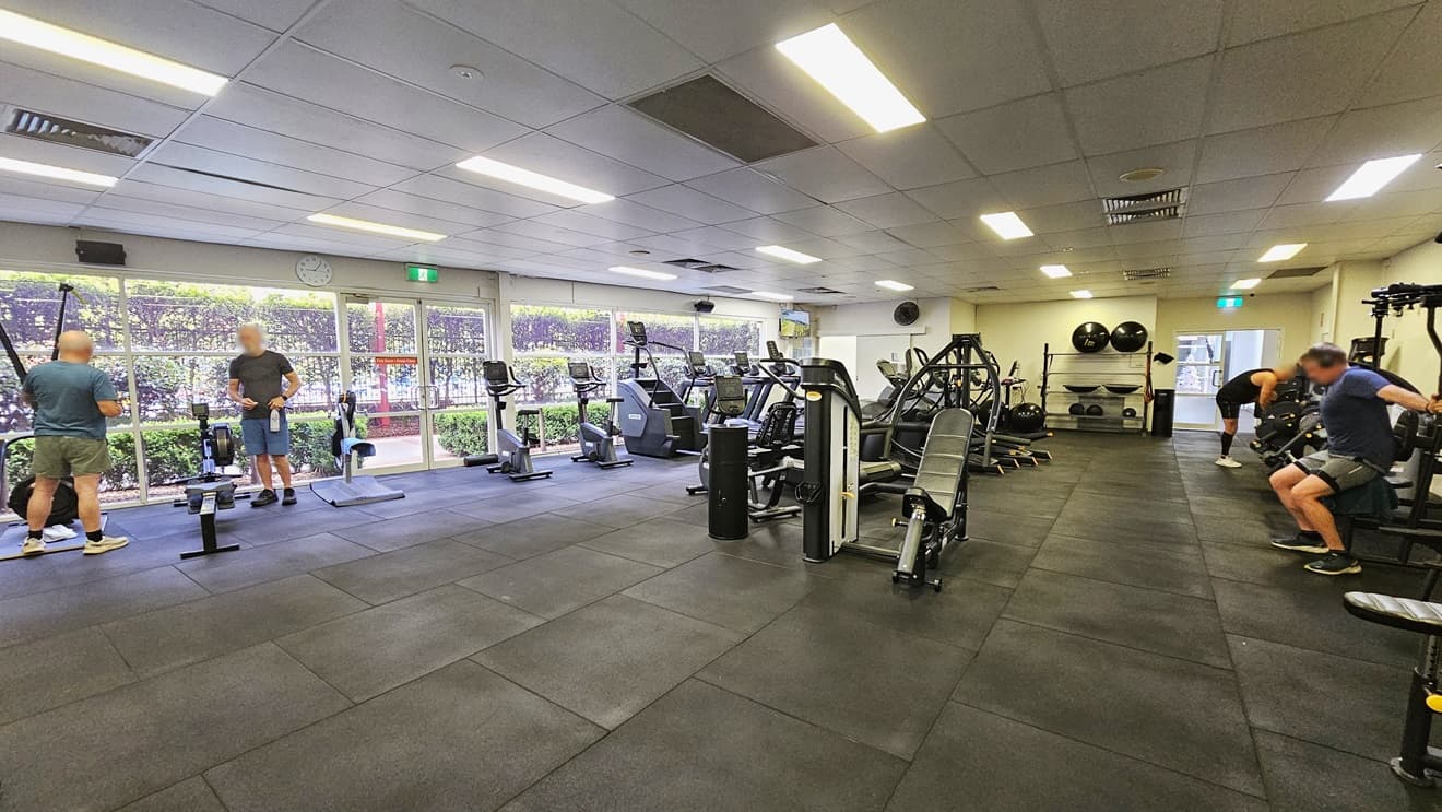 Treadmills and stairmills are positioned near the glass wall on the left. Lateral machines and bench chairs are lined up in the middle, and more strength machines are arranged on the left. Further in, stability balls are displayed. The floor features black rubber tiles, and the ceiling is all white with fluorescent lights. Gym users are visible.