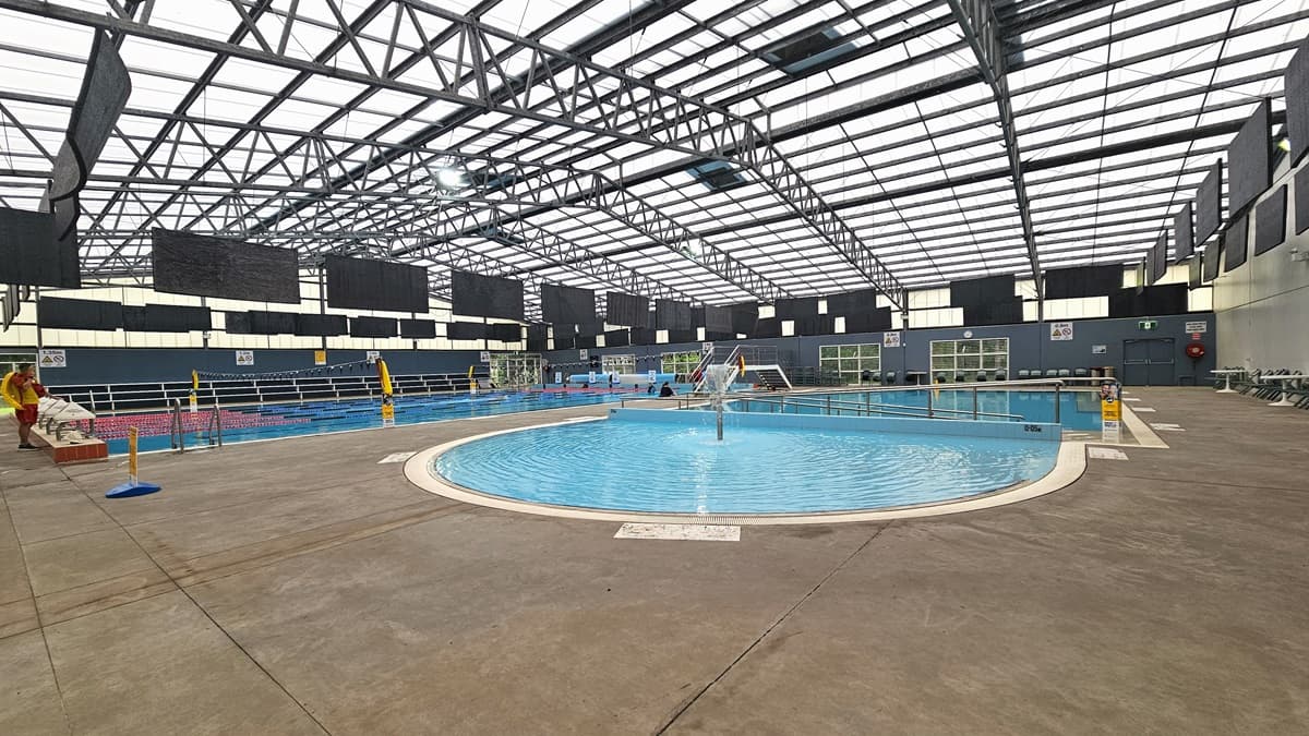 A Christmas tree stands before the glass door. Access to the Indoor Aquatic Centre is through a manual opening glass door equipped with posters. Pots of indoor plants are placed near the entrance. A flat-screen is mounted to the right wall. Inside the Aquatic Centre, the toddler wading pool is directly ahead. The ceiling has an industrial look with exposed galvanized steel beams and translucent roofing.