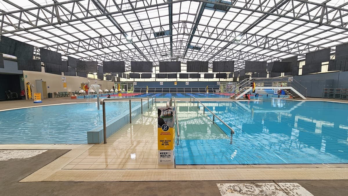 To the left is the toddler wading pool with a fountain, next to it is the activities pool with a ramp entry and handrails. On the far left is the 8-lane, 25m heated lap pool accessed by stairs or the program pool. Tiered seating and plastic chairs line the walls. The industrial-style ceiling has exposed galvanized steel beams and translucent roofing. Black rectangular mats hang from the ceiling, and a safety banner is on the pool deck.