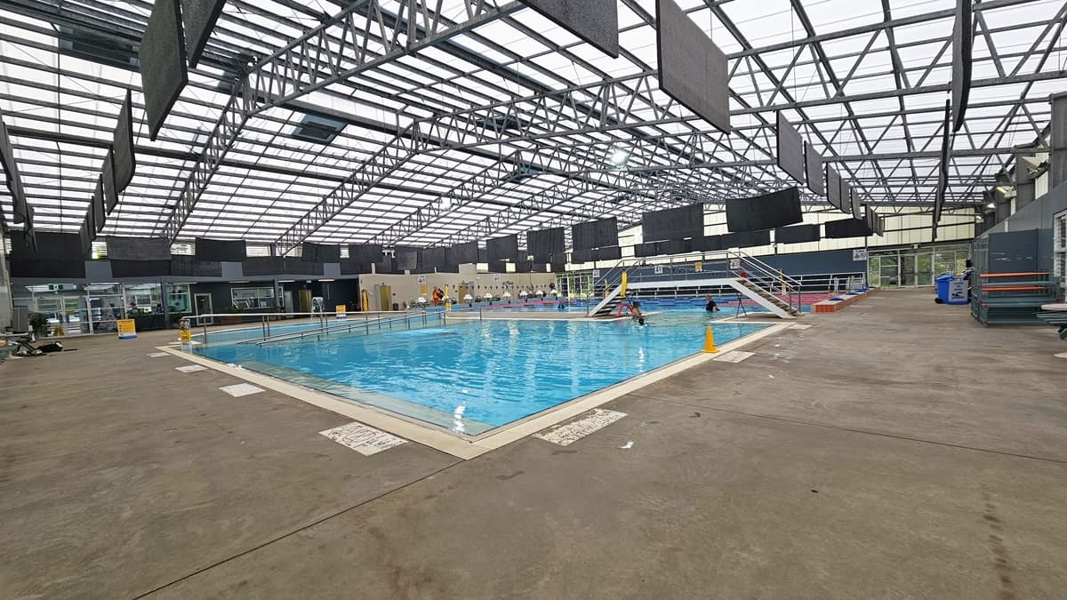 The activities pool features a ramp entry with handrails and stairs with handrails. Safety posters and cones are visible along the pool overflow grid. To the right is a locker area with rubbish bins in place. The industrial-style ceiling has exposed galvanized steel beams and translucent roofing. Black rectangular mats hang from the ceiling, and a safety banner is on the pool deck.