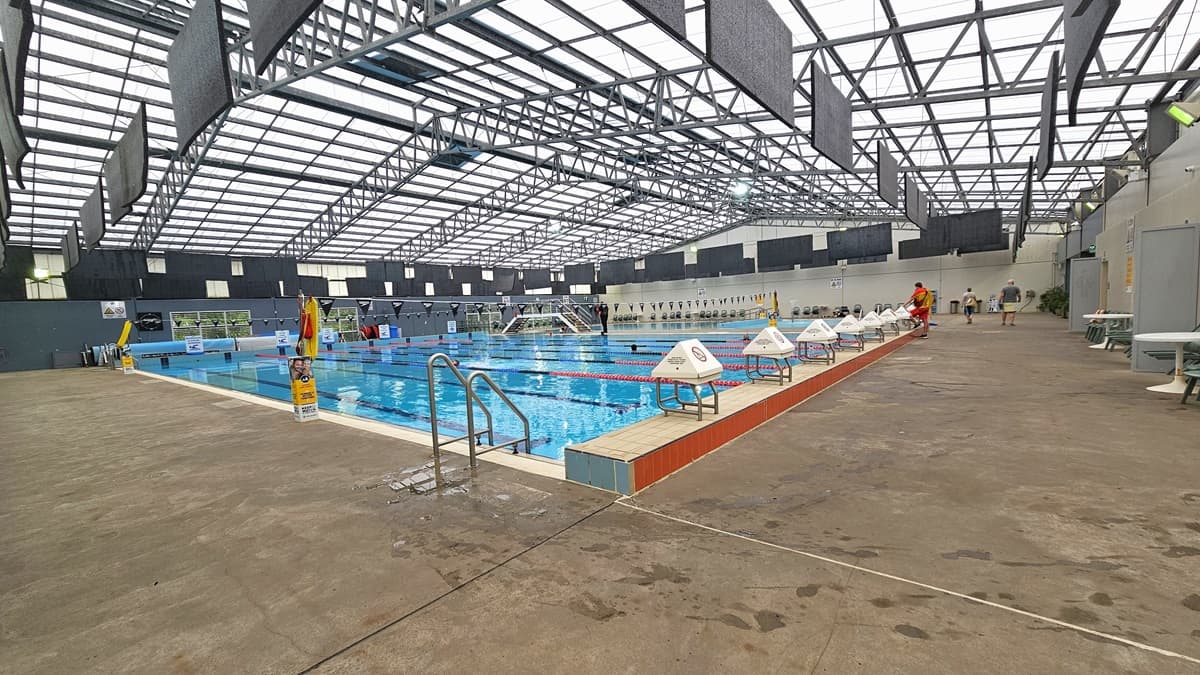 The 8-lane, 25m heated lap pool is accessible via stairs with handrails or through the program pool. Plastic chairs are arranged along the walls on the right. Diving boards with covers are positioned on the raised pool deck. The pool features floating ropes, and a string of black banners runs across it. The industrial-style ceiling has exposed galvanized steel beams and translucent roofing. Black rectangular mats hang from the ceiling, and a safety banner is displayed on the pool deck.