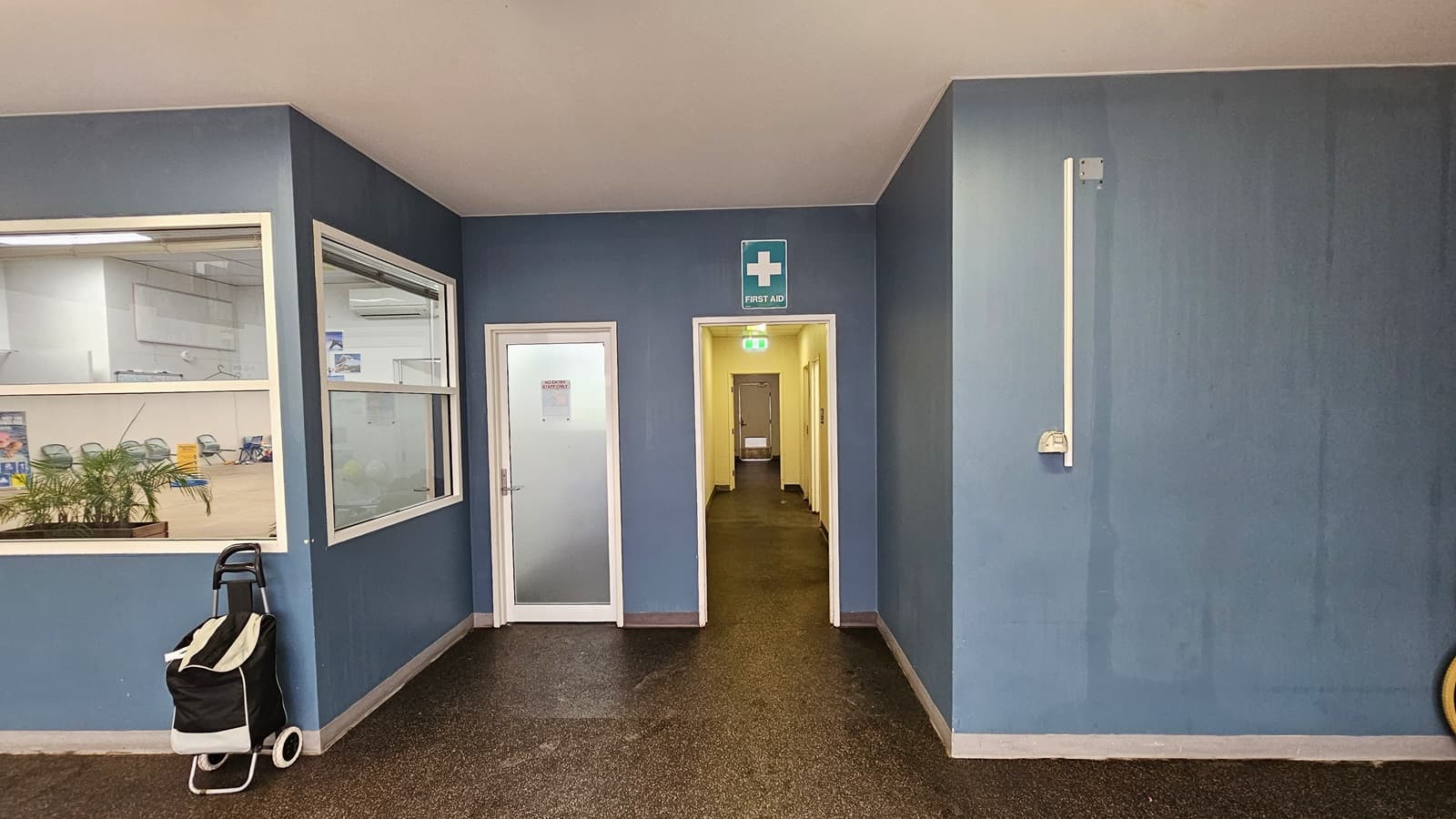 A narrow corridor leads to the accessible bathroom. A "First Aid" sign is affixed to the upper wall. A door on the left opens to a seating area with chairs and a planter box. A trolley bag is positioned against the wall. The floor has grey tiles, the ceiling is white, and the walls are painted blue.