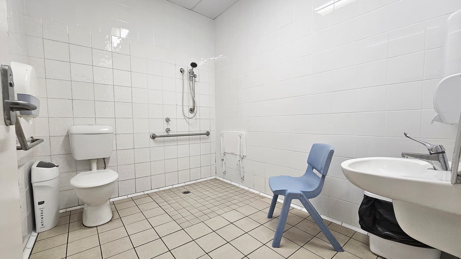Inside the bathroom, a toilet bowl with handrails and a rubbish bin is on the left. Adjacent is the shower with handrails. To the right is a washbasin, a rubbish bin, and a grey plastic chair. On the left, a tissue dispenser is mounted to the wall. The floor has grey tiles, and the ceiling and walls are all white.