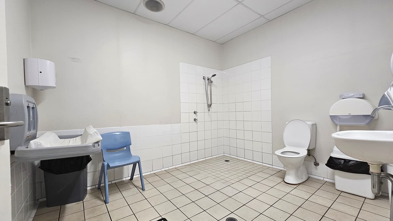Inside the bathroom, a toilet bowl with handrails and a rubbish bin is on the right. Adjacent is the shower with handrails. To the right is a washbasin, a rubbish bin, and a tissue dispenser. On the left, a foldable change table, a tissue dispenser, a grey chair and a rubbish bin are present. The floor has grey tiles, and the ceiling and walls are all white.