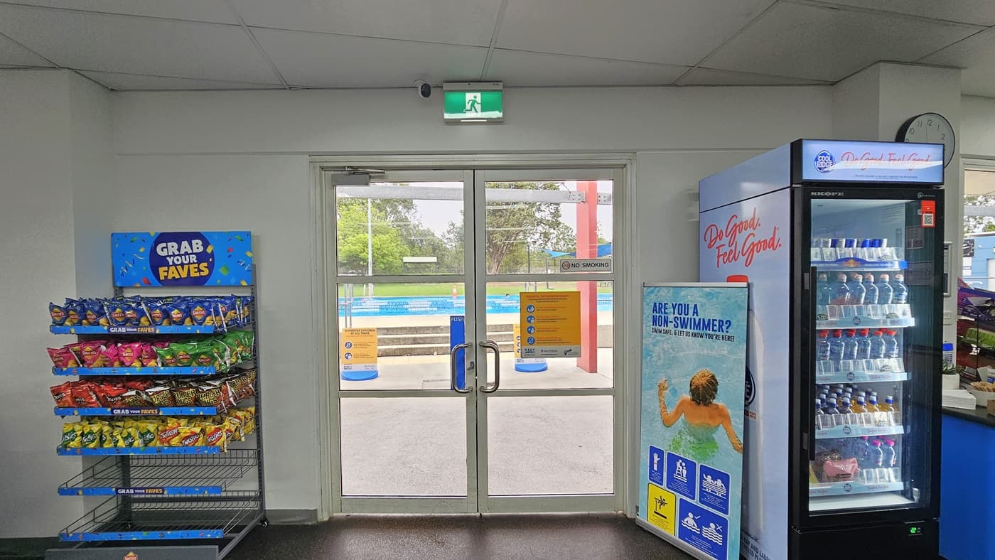Access to the outdoor facility is through a manual glass door adorned with posters and stickers. To the left is a storage rack displaying snacks. To the right is a fridge filled with cold drinks. A clock is mounted on the wall along with an exit sign. A retractable banner stands on the right. Outside, the pool is visible