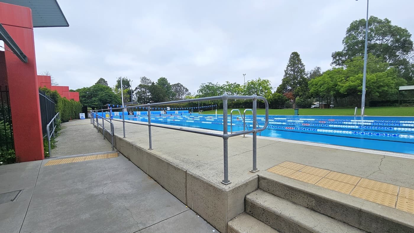 To the left, a ramp entry with handrails and truncated domes. In the middle, steps with handrails and domes. The 50m outdoor pool has steps and handrails on all sides, floating ropes, and yellow banners. Truncated domes are on the floor. On the other side, shade cloths, tables, and chairs under trees.