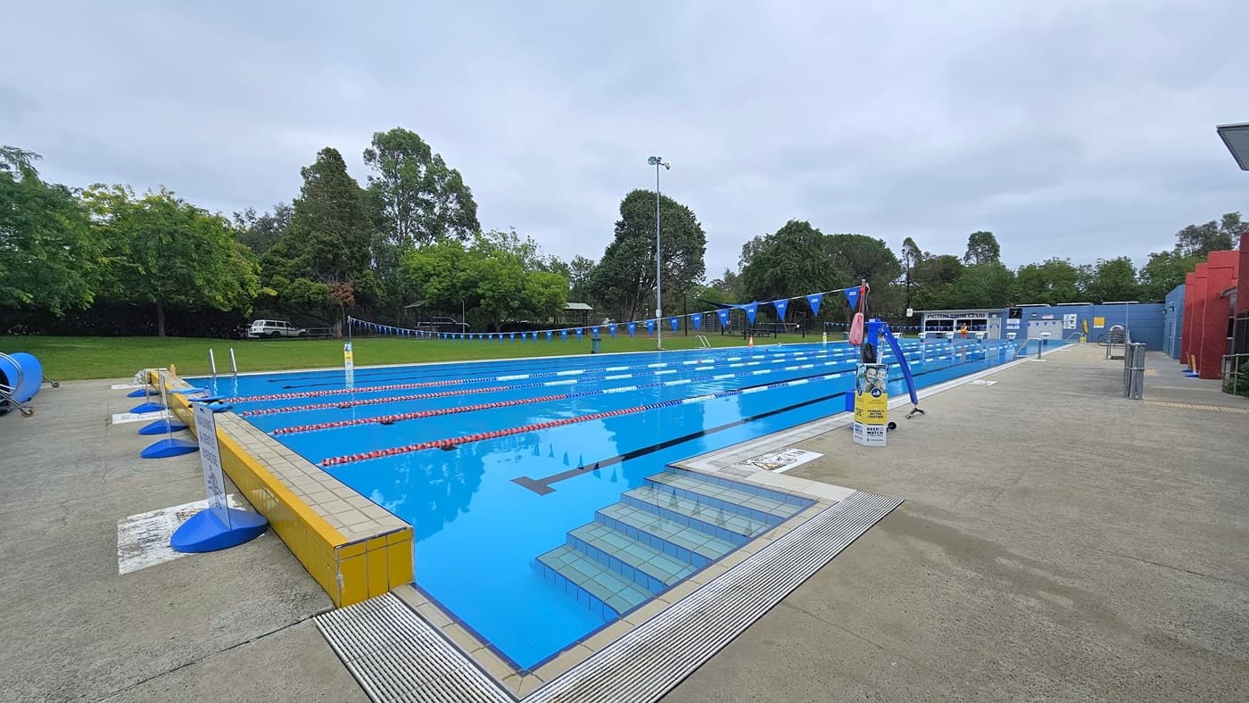 The 50m outdoor pool has a stepped entry with handrails on all sides. Floating ropes and blue buntings run across the pool. Free-standing banners are near the raised pool deck, along with a rolled blue non-slippery mat. On the right, a hoist is positioned near the pool overflow grid. Tall trees and grass surround the area.