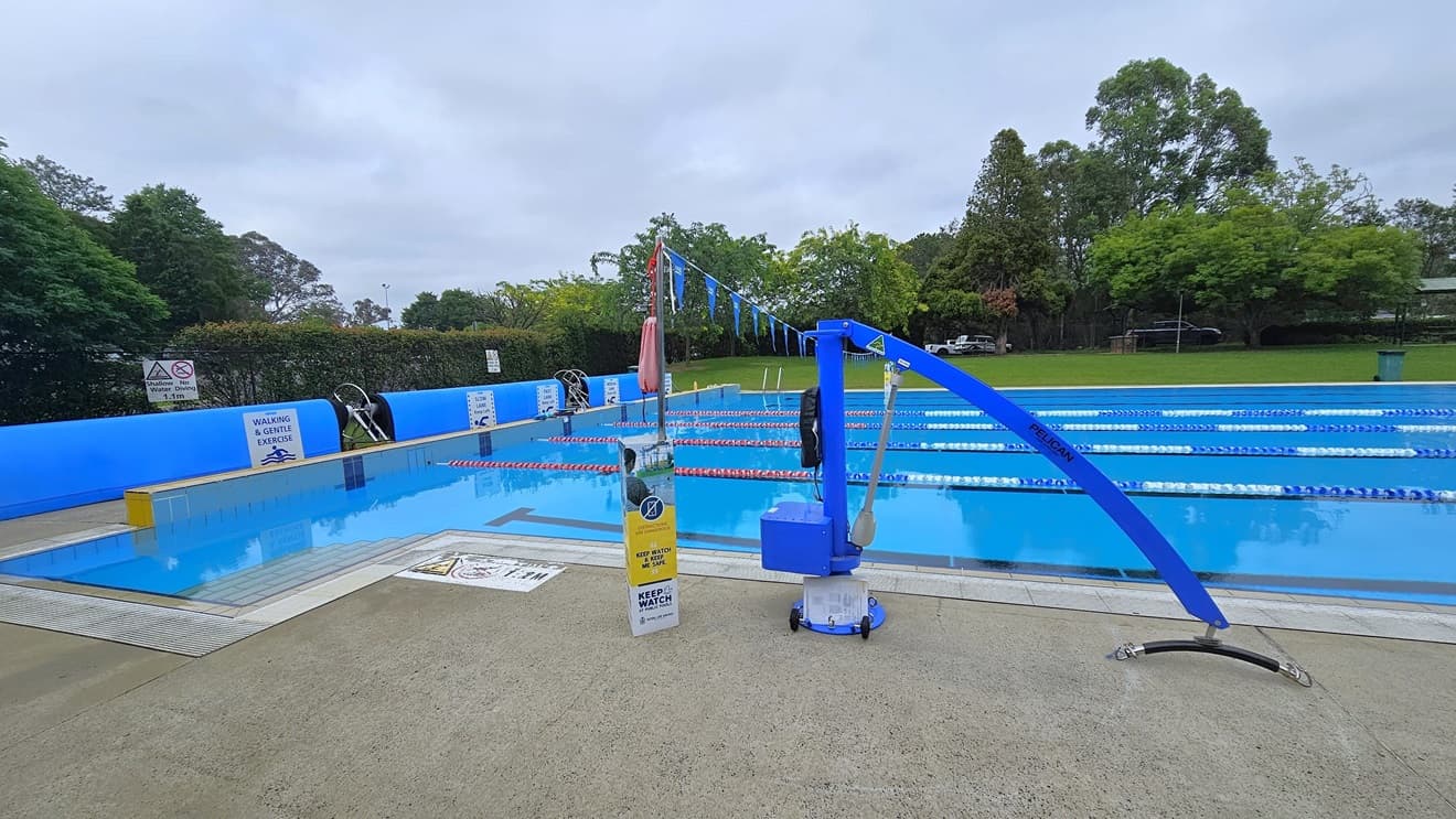 The 50m outdoor pool features a stepped entry with handrails on all sides. Floating ropes and blue buntings run across the pool. On the left, free-standing banners are positioned near the raised pool deck, along with a rolled blue non-slippery mat. A blue hoist is located near the pool overflow grid. Tall trees and grass surround the area.