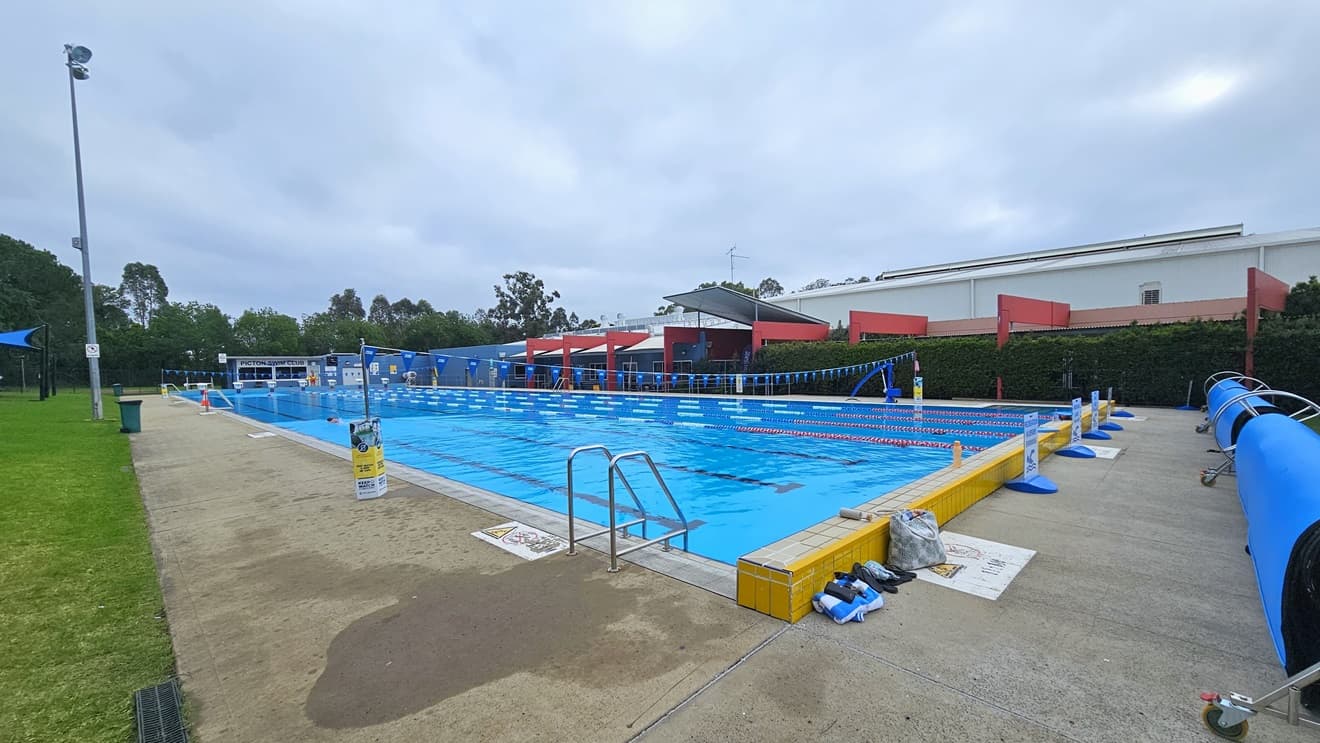 The 50m outdoor pool has a stepped entry with handrails on all sides. Floating ropes and blue buntings run across the pool. On the right, free-standing banners are positioned near the raised pool deck, along with a rolled blue non-slippery mat. A blue hoist is located near the pool overflow grid on the end. Tall trees and grass surround the area.