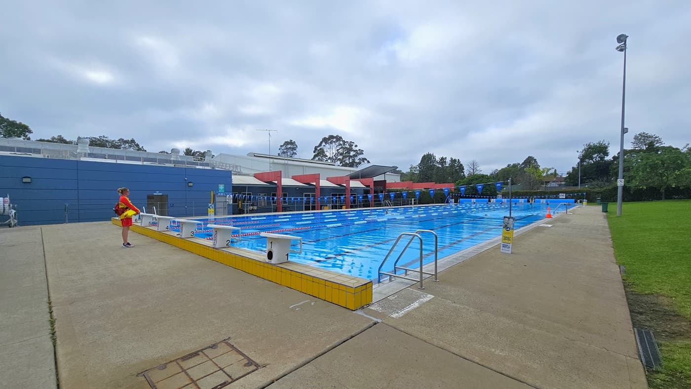 The 50m outdoor pool features a stepped entry with handrails on all sides. Floating ropes and blue buntings run across the pool. Diving boards are positioned on the raised pool deck. Tall trees and grass surround the area. At the end of the pool is the facility building. A pool staff member stands near the pool, and a tall floodlight is positioned on the grass area to the right.