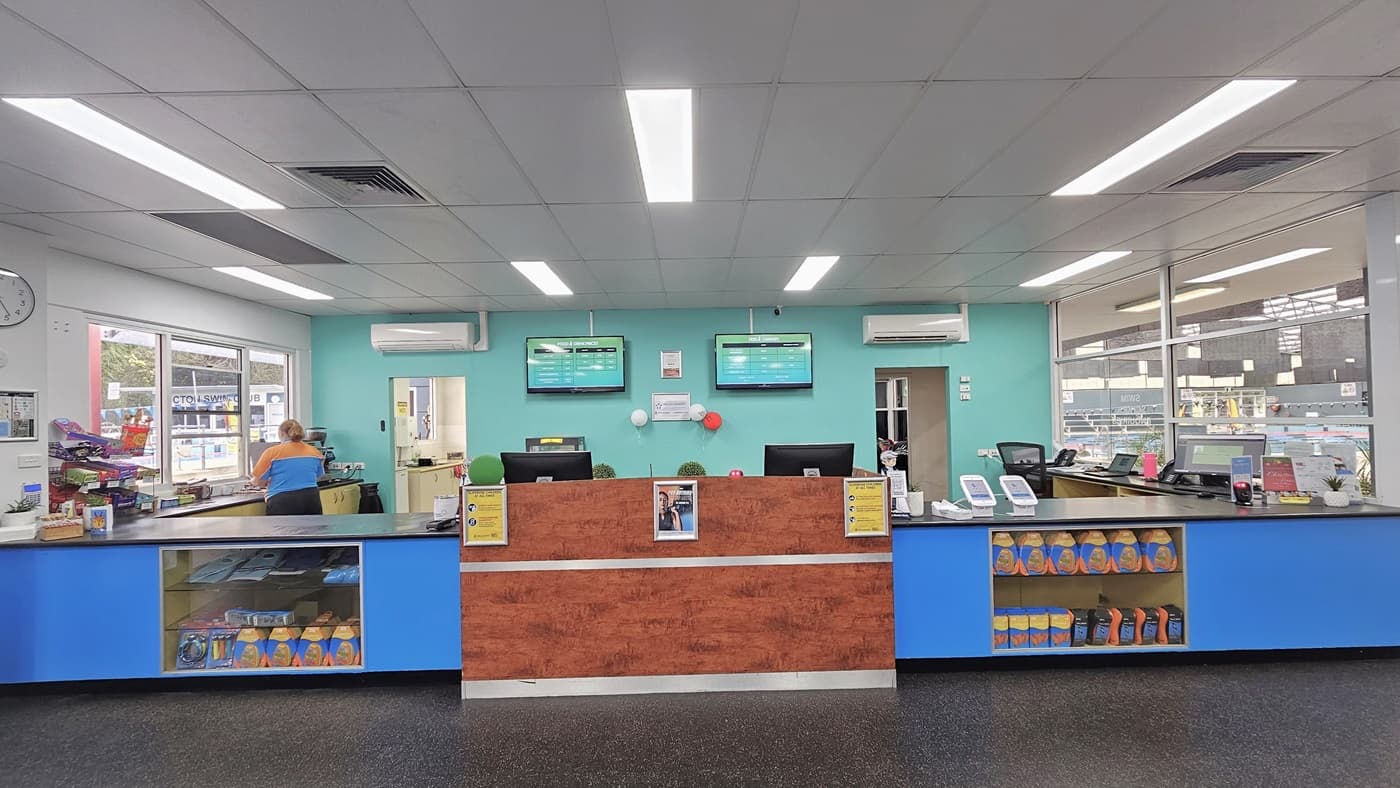 In the center is the reception counter and café, featuring a brown wooden design and a blue countertop with a glass display. Posters are mounted to the front. Swiping machines, computers, and food items are on top of the counter. Flat-screen TVs, air conditioning, and a wall clock are mounted on the wall. The floor has grey tiles, and the ceiling has a white porous design with fluorescent lights.