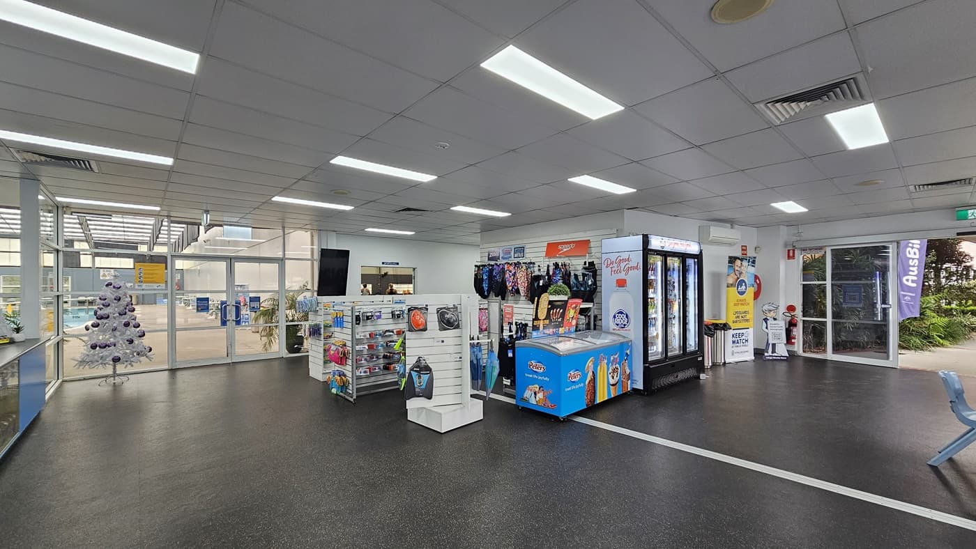 In the center is a retail area displaying swimwear, goggles, and accessories. A fridge with cold drinks is positioned on the right, next to an ice cream freezer. Banners and rubbish bins are placed near the entrance on the right. To the left is a glass door entrance leading to the aquatic center. A Christmas tree stands on the left. The floor has grey tiles, and the ceiling has a white porous design with fluorescent lights.