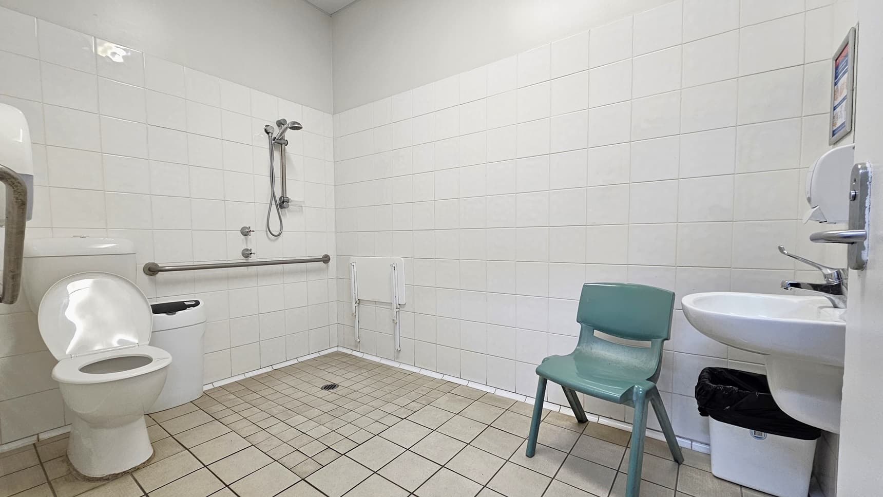 Inside the bathroom, a toilet bowl equipped with handrails and a rubbish bin is positioned on the left. Adjacent to it is the shower with handrails. To the right is a washbasin, a tissue dispenser, and a plastic chair. The floor has grey tiles, and the ceiling and walls are all white.