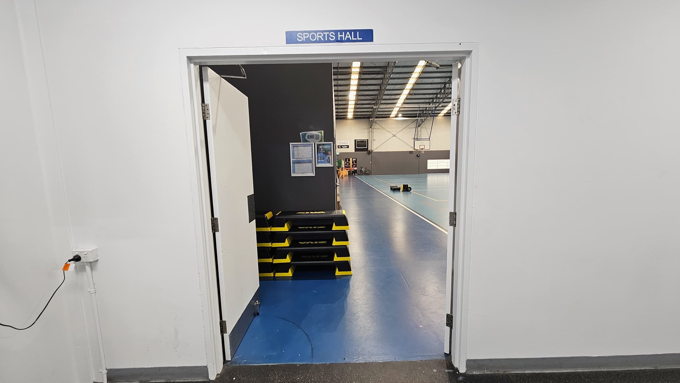 In the middle is a retail area displaying swimwear and accessories. To the right is a glass door leading to the outdoor pool. To the left, a large retractable banner is placed near the wall. Further in, the gym is visible. The floor has black tiles, and the ceiling is white with fluorescent lights.