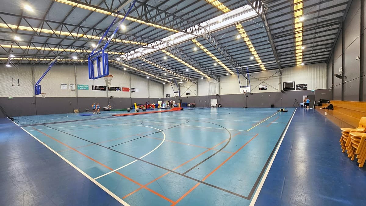 Inside the Sports Hall are two courts, separated by a black mesh curtain suspended from the ceiling. The floor is in a contrasting blue colour, marked for various ball games. The ceiling has an industrial look, featuring exposed metal beams with lines of polycarbonate corrugated roofing. Tiered timber seating is positioned on the left along with yellow plastic chairs. The walls are adorned with large posters. A few people engage in an activity on the far end.