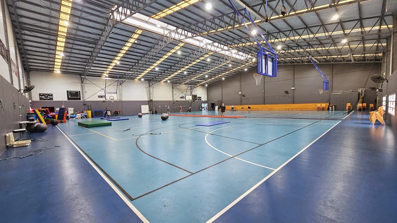 Inside the Sports Hall are two courts, separated by a black mesh curtain suspended from the ceiling. The floor is in a contrasting blue colour, marked for various ball games. The ceiling has an industrial look, featuring exposed metal beams with lines of polycarbonate corrugated roofing. Tiered timber seating is positioned on the other side along with yellow plastic chairs. The walls are adorned with large posters. A few people engage in an activity in the left corner.