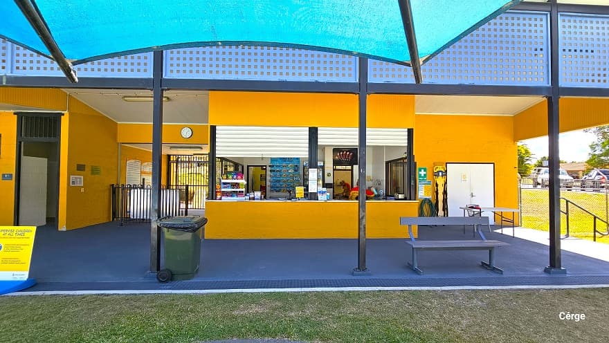 Image shows the front of the low-set administrative building with orange brick walls and dark grey roofing. The reception area can be distinguished by the blue perforated rectangular section that covers the ceiling and a stair with metal handrails on the right side leading to the toddler pools. The middle entryway connects to the concrete pathway and the pool deck. In front of the buildings are two blue sun shades with a large grey circular floor area within a grass section.