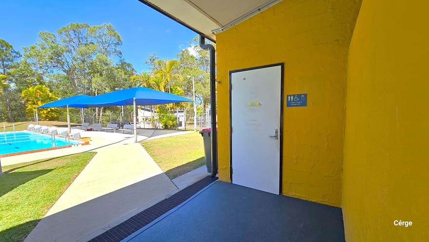 Accessible bathroom at Woodform Swimming Pool. It has a white door with black outline on the edges. On the left wall is a black gutter that drains directly to the black grating channels for water drainage. On the orange brick wall on the right is a blue sign for ‘Unisex toilet and shower’. This front area of the building has a grey floor. Seen from this position is the seating area beside the 6-lane outdoor pool.