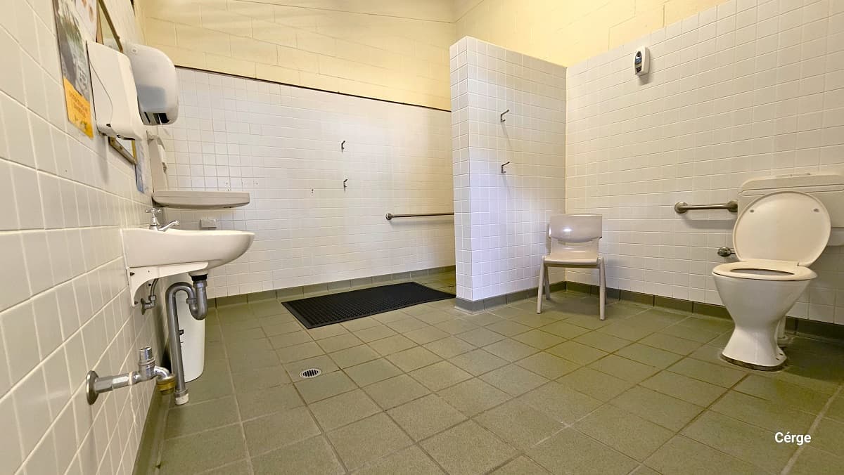 Inside the accessible bathroom at Woodford Community Swimming Pool. The room has a grey tile floor, small white square tiles, and a cream brick wall. On the right end is the toilet, a light brown plastic chair, and grab bars. Directly in front is the wash basin, liquid soap dispenser, toilet roll holder, and a baby changing table. A white tiled wall separates the shower and toilet section with black grating floor channels for water drainage.