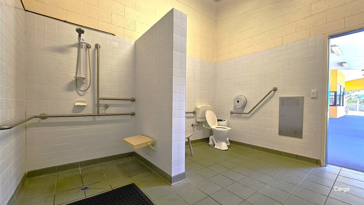 Closer look at the shower section inside the accessible bathroom. Next to the shower head is a grab bar that forms an inverted T on the white tiled wall to aid persons with mobility disabilities. Mounted on the wall to the right is a small, cream, shower seat. This photo angle also shows a grab bar on the left side of the toilet. There is a rectangular metal next to a white switch beside the door.