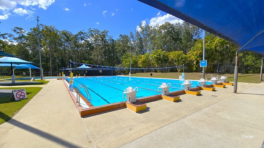 Starting section of the 6-lane outdoor pool at Woodform Swimming Pool. The starting section has white diving blocks with side handles placed on top of the elevated orange tile floor section with yellow sides. Each lane is separated by red, blue, and white floating rope lanes. On the sides are stainless steel pool ladders. On the left side, within the grass section, is a white ‘No Diving’ sign. Blue sun shades can also be accessed on the left side.