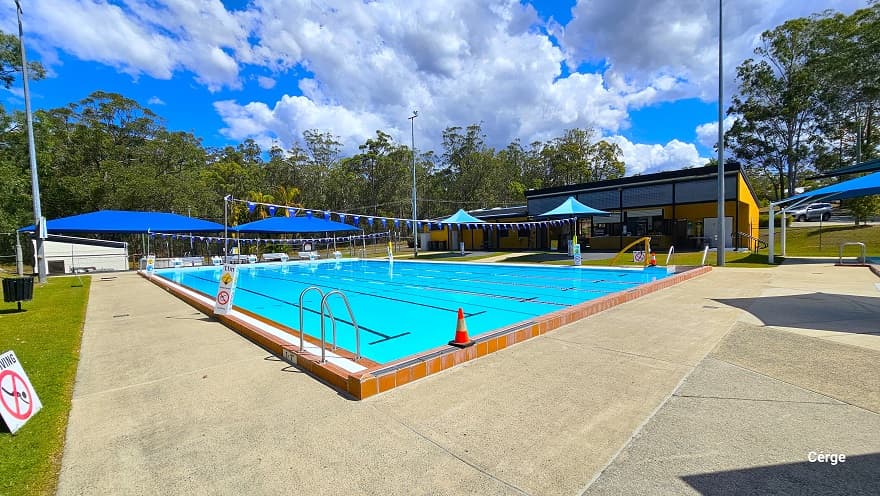 A closer look at the 6-lane outdoor pool when standing near the white ‘No Diving’ sign. There is an orange cone on top of the orange tile on the side of the swimming pool. An adequate pool deck allows guests to walk around the pool. On the opposite side of the pool is an orange pool hoist. Hanging above the pool are strings of blue triangular flags. Within the pool ladder is a small section with a white tile floor in between orange tiles.