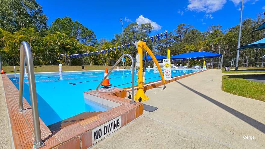 Orange stair tile entrance next to the yellow pool hoist. On the sides are metal handrails to aid people while entering the pool. A white “No Diving” sign is placed on the top of the stairs. Another orange cone can be found between the pool stairs and the pool hoist. A white rectangular narrow sign for 1.1m depth is placed on the right pool edge. Wrap around the pool flag post is a yellow floating device.