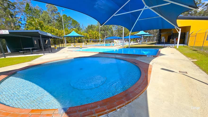 Shallow circular and rectangular pool for toddlers right next to the dark grey corrugated iron shed for storage. Both pools have red tile edges. On the side of the roof are white “No Diving” signs. There is a picnic-style grey table within the dark grey corrugated iron shed. A blue sunshade with white metal frames offers shade to both pools.