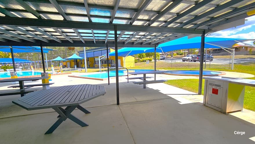 Seating section of the dark grey corrugated iron shed. The ceiling has silver padding and dark grey metal beams. There are wooden picnic-style tables and an octagon-shaped green table. On the right side is a stainless steel electric BBQ. This section offers a view of the carpark outside and all the pools inside the facility. The surrounding gum trees give a tranquil atmosphere to Woodford Swimming Pool.