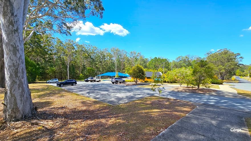 Concrete pathway leading to the dedicated carpark with a few parked cars at Woodford Swimming Pool. It has white markings on the floor for each car park space. There are medium-tall trees planted within a circular garden plot within the carpark. Further to the left is a large blue sunshade and a low-set orange building with corrugated grey roofing. On the right is the main road.