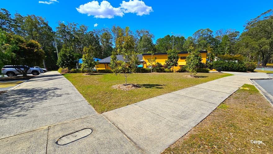 Arrival at Woodford Swimming Pool is through the concrete pathway connected from the main road. The pathway is composed of granite floor square blocks with rough surfaces. There is a large green lawn with newly planted trees before this pathway, and it is next to the low-set yellow building with a dark corrugated roof. Bushes and shrubs are placed aesthetically around the facility.
