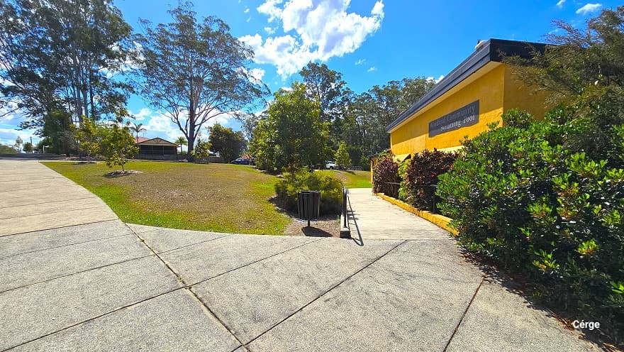 The concrete pathway connects to another concrete ramp with handrails on the side of the yellow-brick, low-set building with a dark grey corrugated roof. The concrete pathway is composed of granite floor square blocks with rough surfaces. On the upper right wall of the building is the Woodford Community Swimming Pool, detailed in white paint over a dark grey background. The residential houses to the back are evident from this position.
