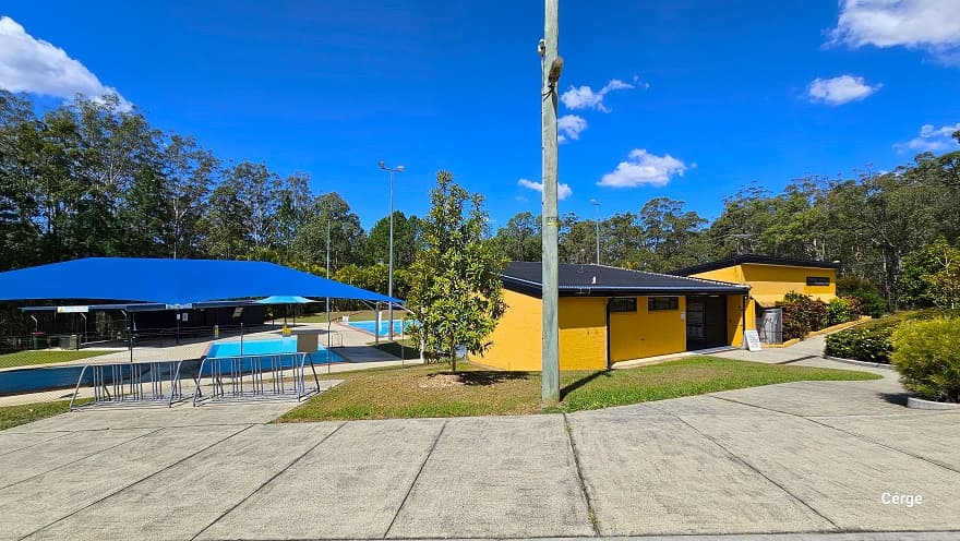 Concrete pathway near the two small toddler-wading outdoor pools and the administrative center with orange brick wall and dark grey corrugated roofing. On the pathway near the pool are two metal barriers. Beside it is a grass terrain with a medium-tall tree and a lamp post. A large blue sunshade covers the toddler pool. This image also shows the entrance to the orange brick admin building on the right side with two small windows on the upper left wall.