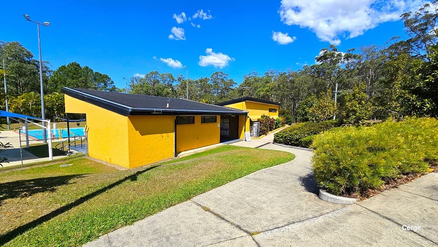 Downward inclination of the concrete pathway leading to the entrance of the administrative center with orange brick wall and dark grey corrugated roofing. The left side of the concrete pathway is a grass terrain, while on the right side is a garden with hip-high shrubs and bushes maintained at the same height. There is a white signboard in the entryway. A black chain-linked fence secures the outdoor pools.