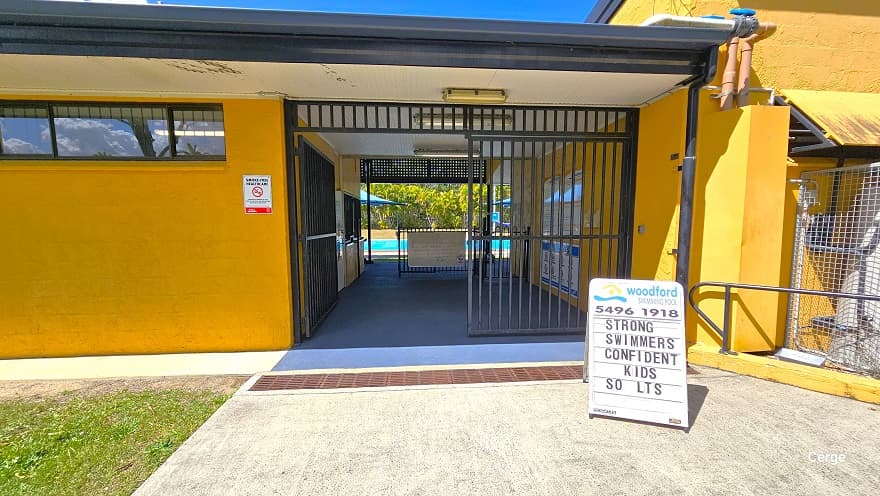 Entry to the administrative center with orange brick wall and dark grey corrugated roofing is through a black gate. On the upper left wall is a small white sign for no smoking. The telephone number of Woodford Community Swimming Pool can be found on the white signboard just before the brown water drain. The building has a blue floor and a white corrugated ceiling.