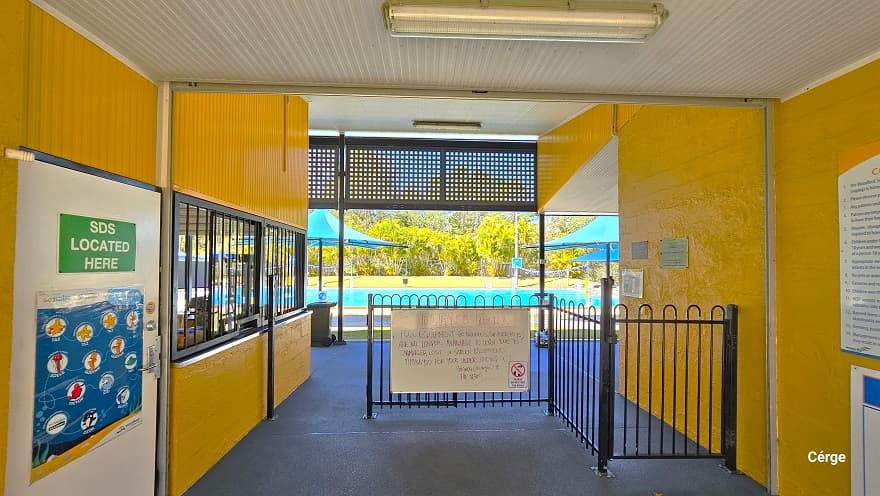 Reception of Woodford Swimming Pool. There is a white door with blue graphics showing different animated sea creatures. Above the door is a green sign for ‘SDS Located Here.’ A low iron black fence with curvy designs regulates access to the pools. Hanging on the iron black fence is a large whiteboard with a write-up to remind guests of the dos and don’ts. Black iron frames protect the reception glass windows. A black perforated rectangular section covers the end of the ceiling.