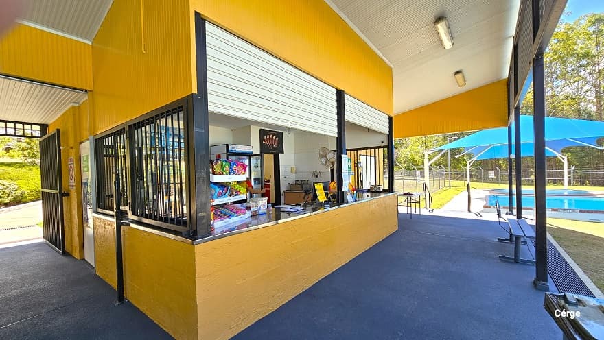 Reception at Woodford Community Swimming Pool also offers snacks and drinks. The reception has a silver countertop and aluminum roller shutters. The lower wall is made of orange brick wall while the upper wall is made of corrugated orange wall. Found on the white corrugated ceiling are rectangular artificial lights. The floor in front is grey with grey metal benches. Covering the toddler-wading pools to the right of the reception is a blue sunshade with white metal supports.
