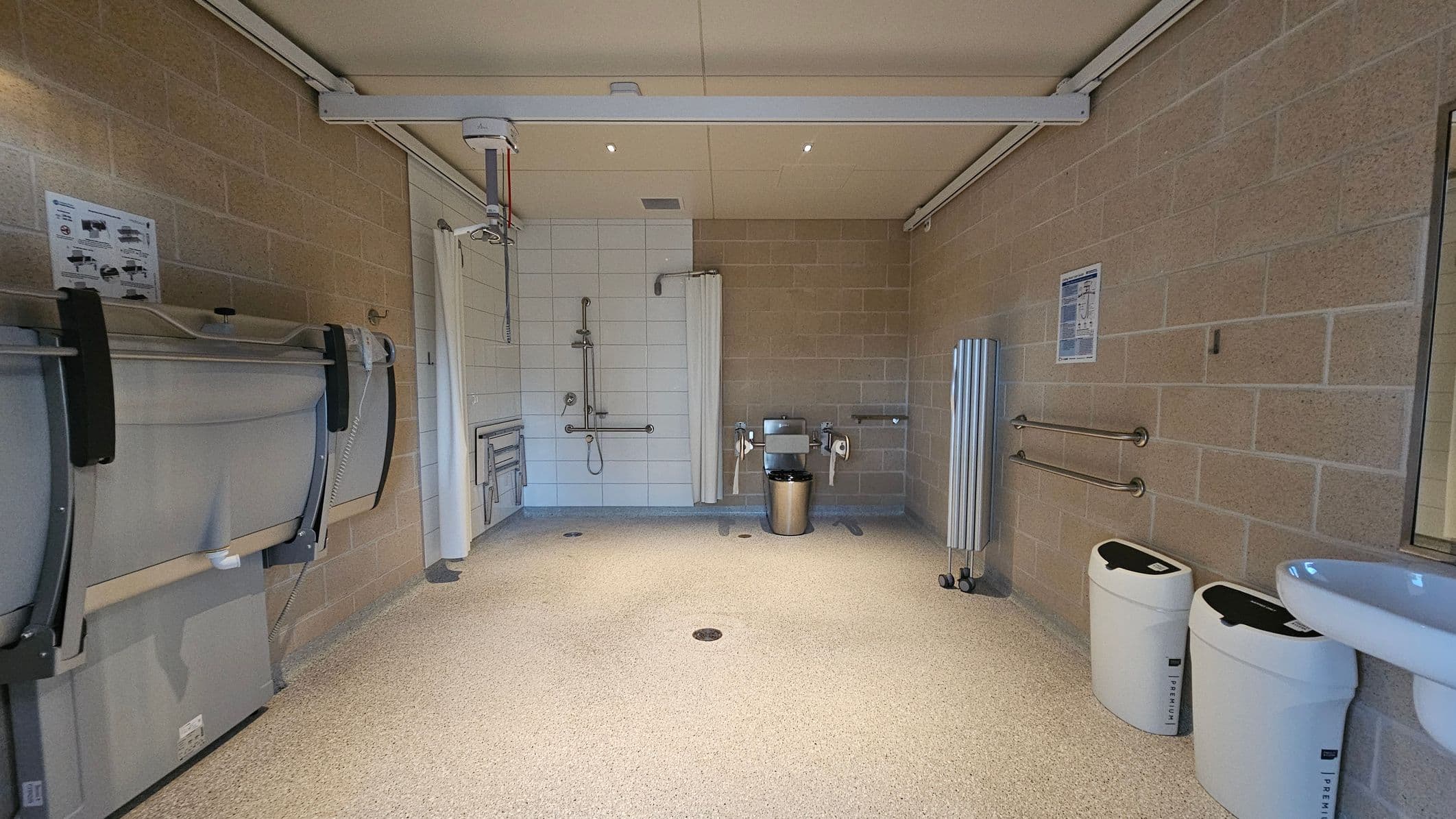 A spacious, accessible bathroom with beige tiled walls and a speckled floor. On the left, there's a foldable changing table and a ceiling track hoist. In the centre, a shower area with a curtain and grab bars. On the right, a toilet with support rails and a bin nearby. Two white bins are positioned against the right wall, and a sink is partially visible. The ceiling is plain with recessed lighting.