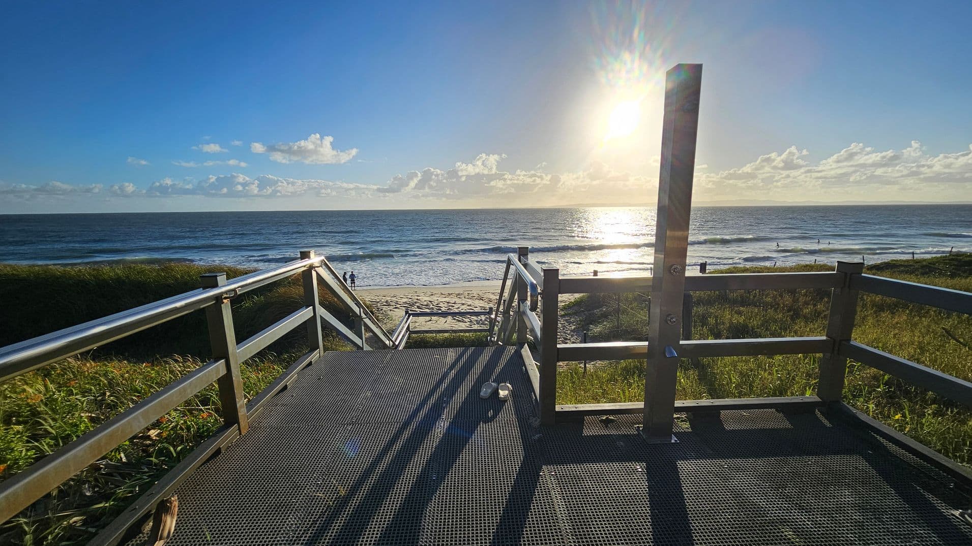 A metal walkway with railings leads down to a sandy beach. The sun is low in the sky, casting long shadows on the walkway. On the left and right, grassy dunes border the path. In the distance, the ocean stretches out with gentle waves, and a few people are visible near the water's edge. A pair of shoes is placed on the walkway.
